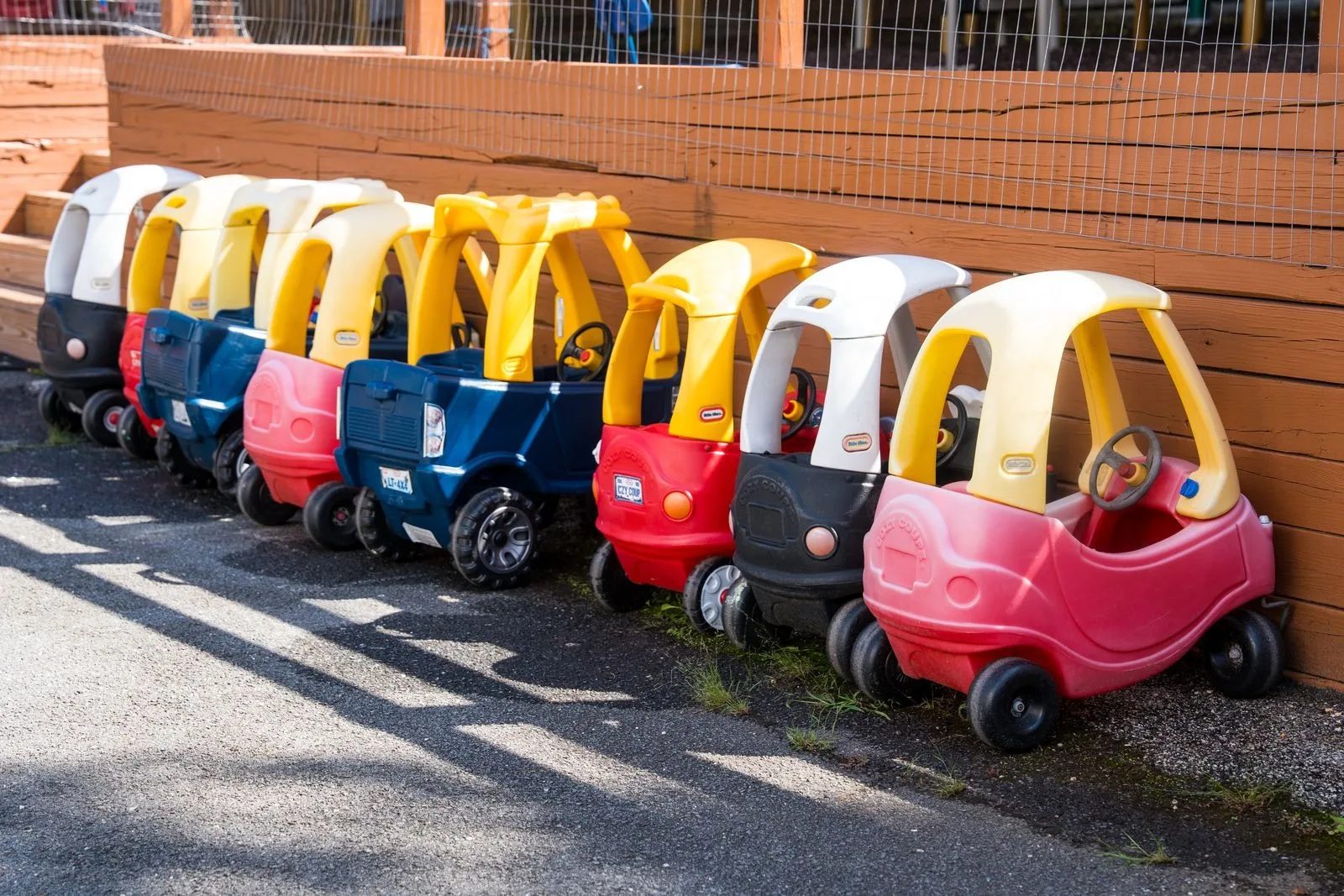 A row of ride-on cars parked along the play yard fence